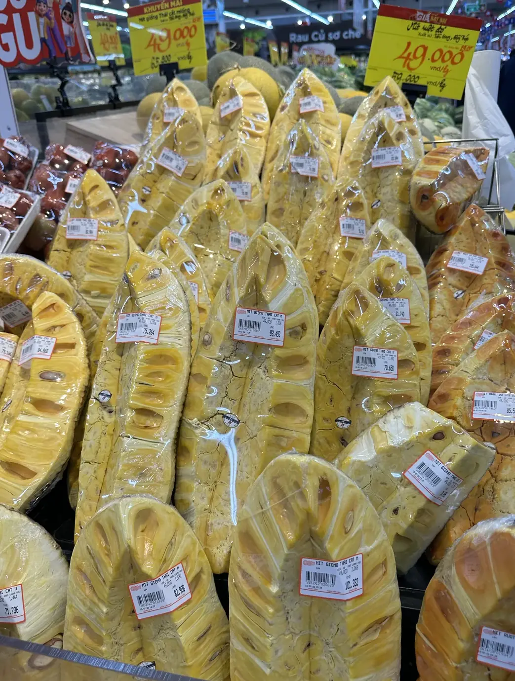 スーパーの青果売り場に並ぶジャックフルーツのむき身。「49.000 VND/kg」の特売価格を示すポップが掲げられ、個々のパックには重量に応じた値札が付いている実写真